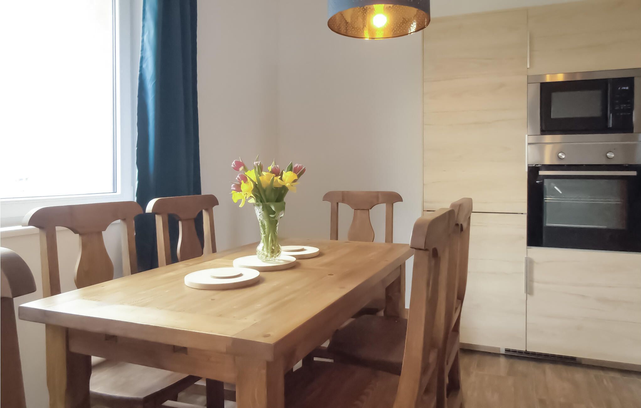 Uma mesa de jantar com cadeiras, um vaso de flores e uma cozinha moderna com eletrodomésticos.