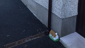 Rakennuksessa on kuvioitu ulkopinta, ikkunassa on valkoinen verho, ovessa on messinkinen kahva ja maassa on pieni pienoismallitalo.