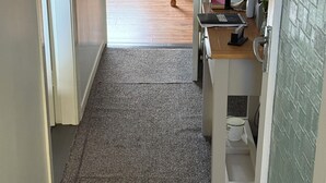 Matolla päällystetty käytävä johtaa huoneeseen, jossa on sänky, pöytä ja lamppu.