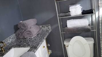 A bathroom with a white sink, marble countertop, and a toilet with a white lid. There are towels and toiletries on shelves.