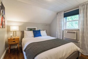 A bedroom with a bed, a bedside table with a lamp, a window with curtains, and an air conditioning unit. - Charming Hideaway in Westport | 16 (KCMO)