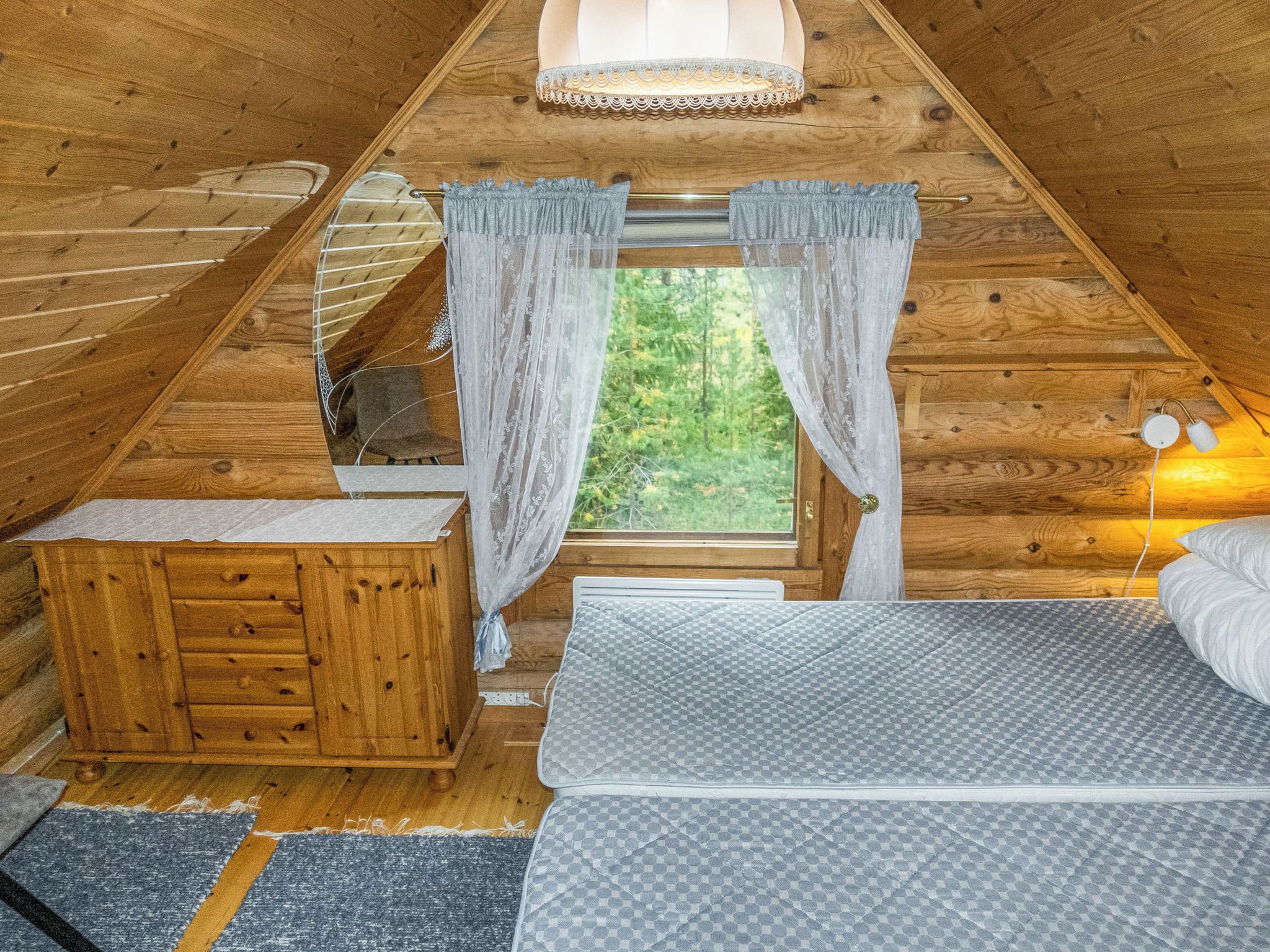 Un dormitorio acogedor en el ático con techo inclinado de madera, una cama con colchón estampado, un aparador de madera y una ventana con cortinas traslúcidas.