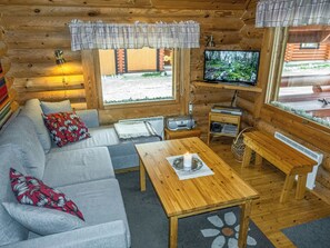 A cozy cabin interior with a sofa, a wooden table, a television, and a window overlooking a forest.