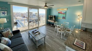 A living room with a sofa, coffee table, TV, and dining area with chairs. There is a large window with a view of buildings and a beach. - Find your beach bliss! (San Diego)