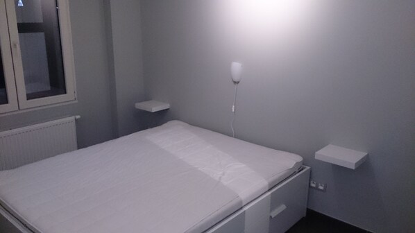 Een kleine eenpersoonskamer met een wit matras, een nachtkastje en een wandlamp.
