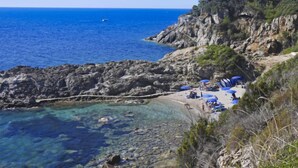 A rocky coastline with clear blue waters, a small beach with umbrellas, and a path leading down to the water. - Mimose 2 by Interhome (Rio Marina)
