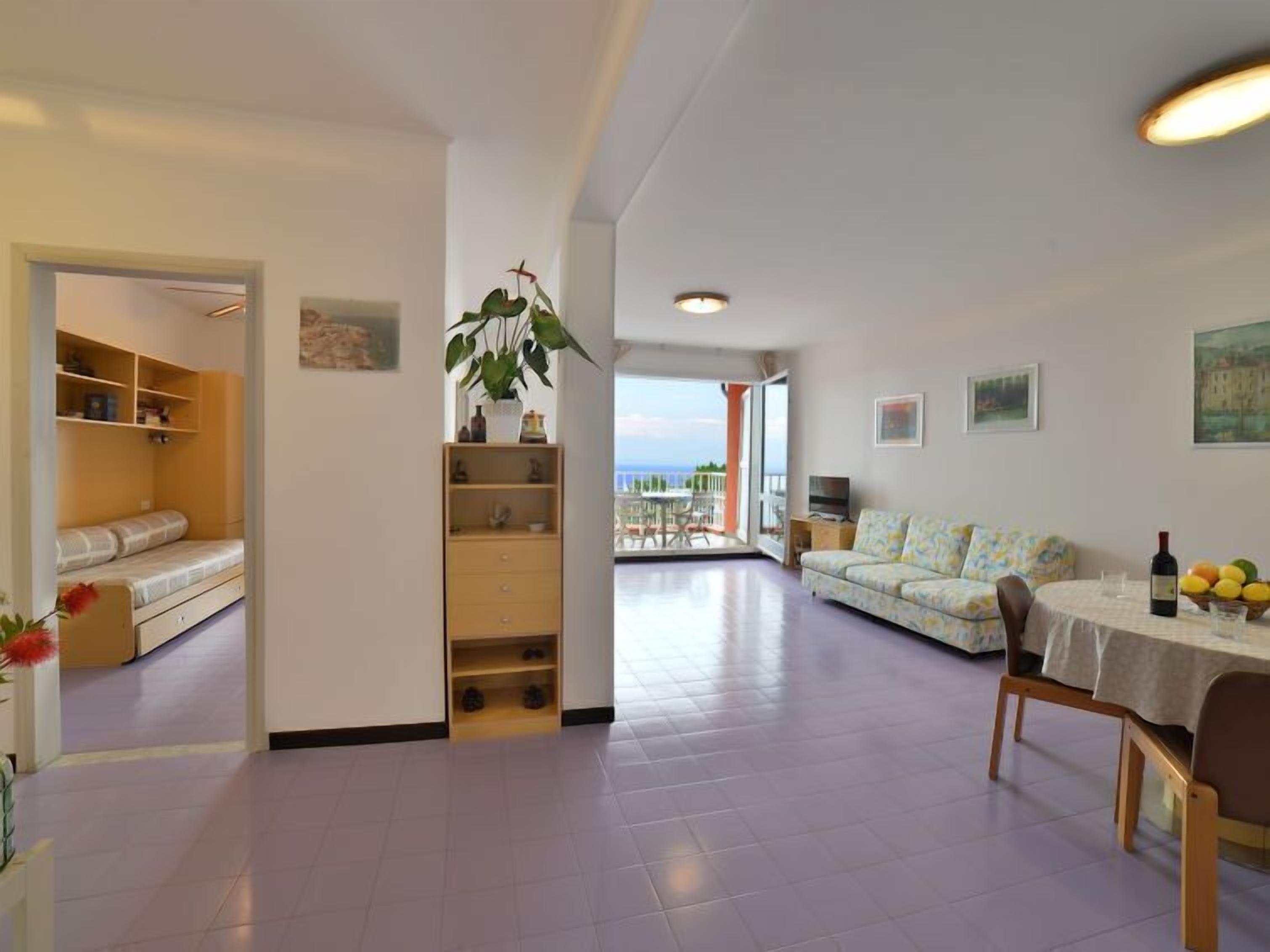 A spacious living area with a sofa, a dining table with chairs, a bookshelf, and a balcony with a view.