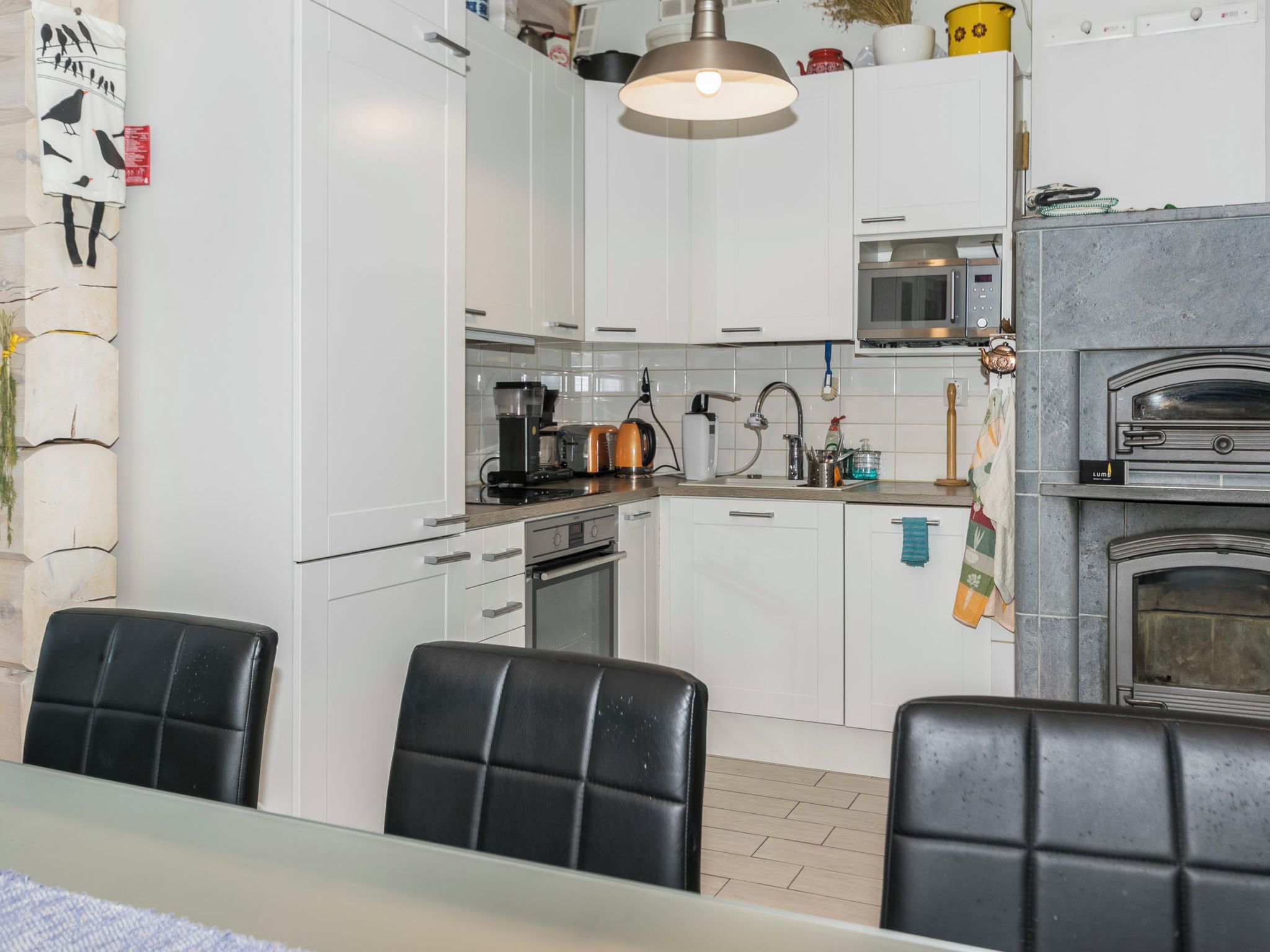 A modern kitchen with white cabinets, a built-in oven, a microwave, and a pizza oven. There are black chairs around a table, and a hanging light fixture above.