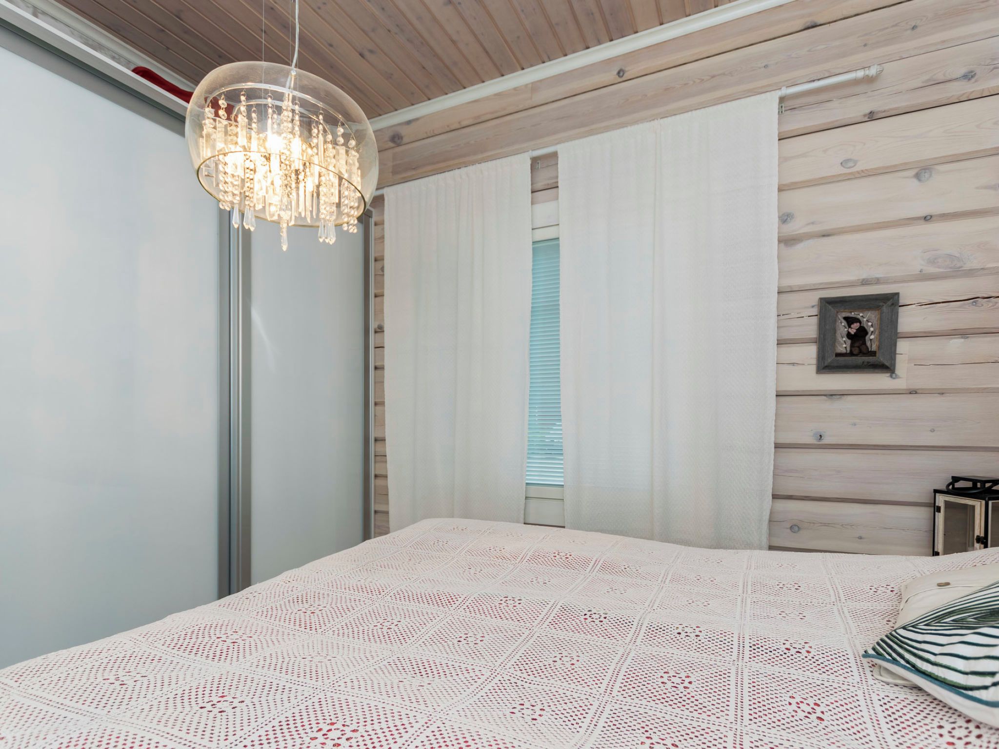 A bedroom with a large bed, a chandelier, wooden panel walls, and a window with blinds.