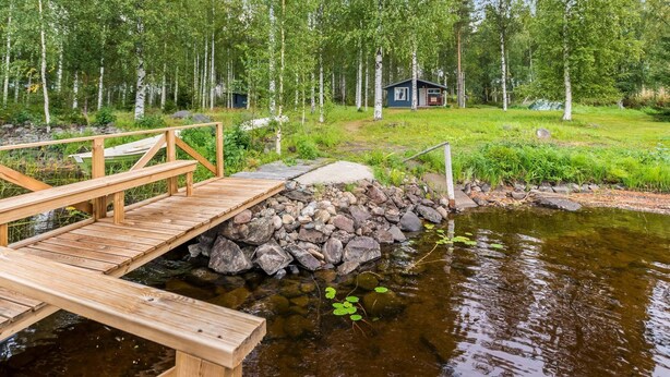 A wooden deck over a rocky stream with a small house in the background surrounded by birch trees.