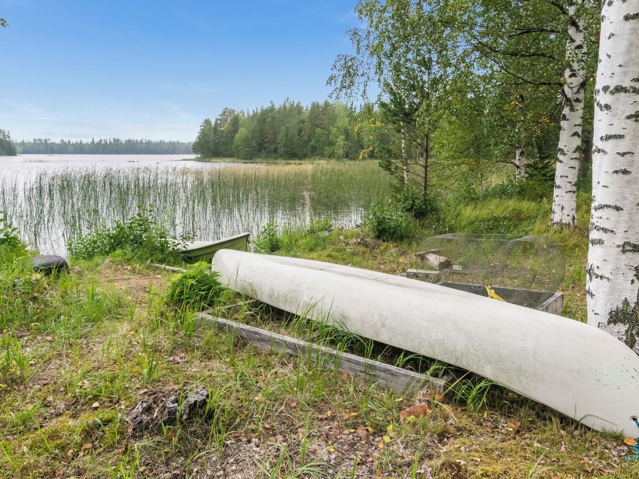 Maisema järven rannalla, jossa betonista valmistettu kanootti lepää rannalla. Sen ympärillä on ruohoa, puita ja kirkas taivas.