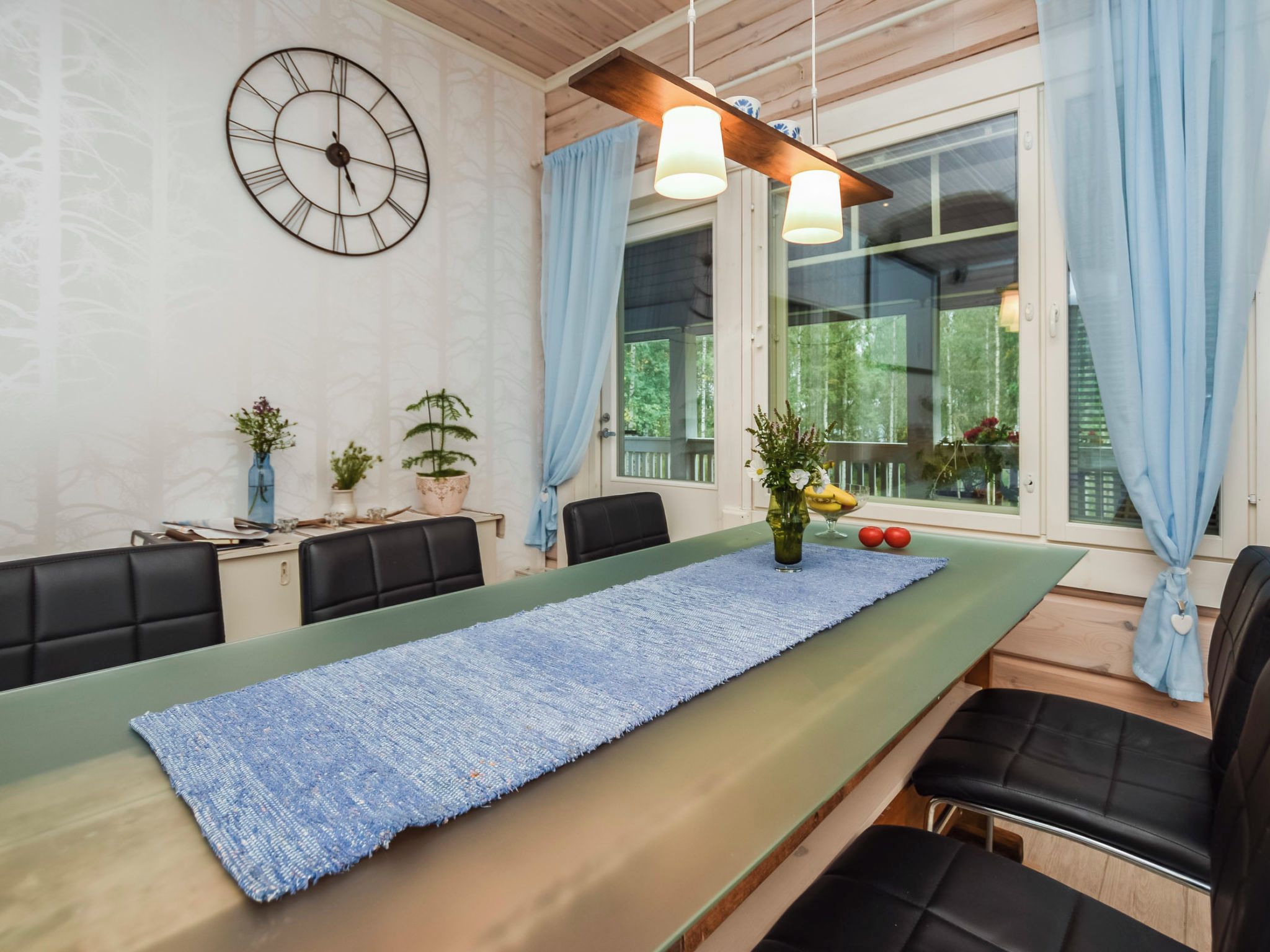 A dining area with a long table, chairs, a clock, and a window with curtains.