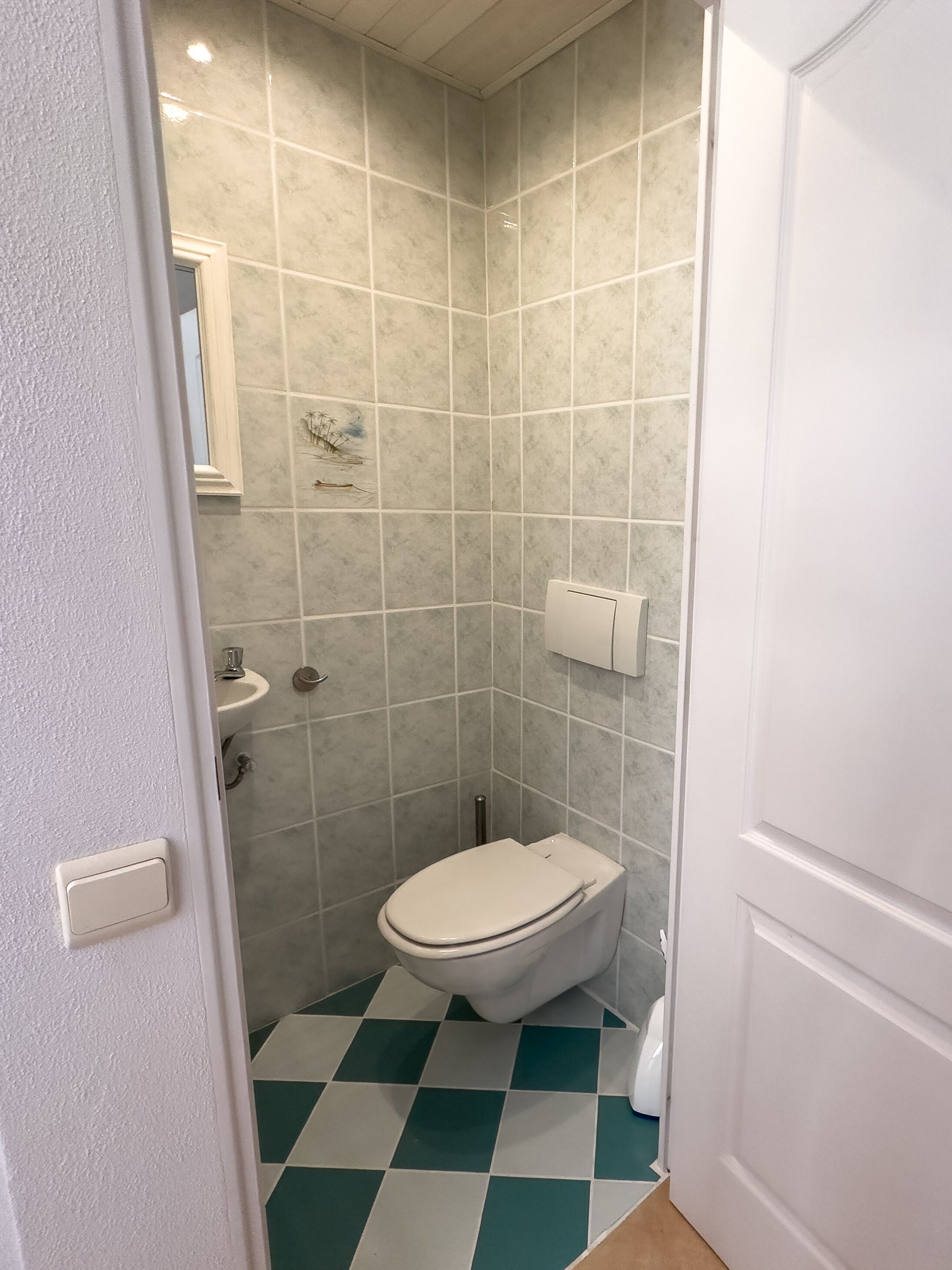 A bathroom with a white toilet, a sink, and a mirror. The walls are tiled, and the floor has a checkered pattern.