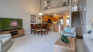 A spacious living area with a dining table, chairs, a sofa, a TV, and a staircase leading to an upper level. - Ocean-View 2BR Maui Condo with Lanai, Steps from the Beach (Kihei)