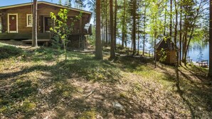 En hytte med terrasse og mindre udhus ved en sø omgivet af træer.
