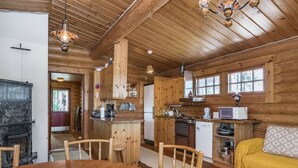 Indbydende interiør i en hytte med et træbord, stole, sofa, pejs, et køkken med hårde hvidevarer og et vindue.