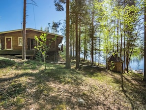 A cabin with a deck and a small shed, surrounded by tall trees and a body of water.