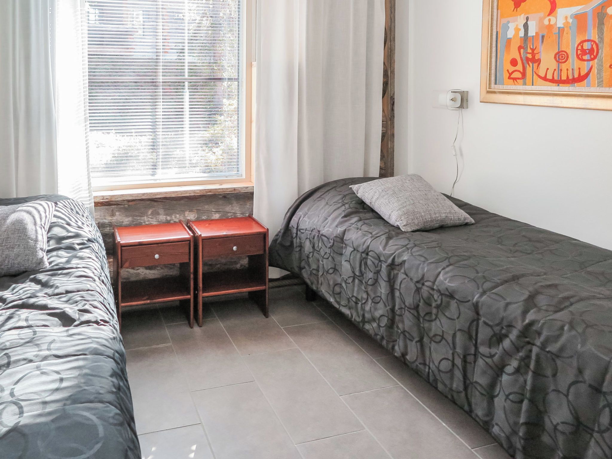 A bedroom with two single beds, a wooden nightstand, a window with blinds, and a wall-mounted power outlet.