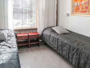 Une chambre avec deux lits simples, une table de chevet en bois, une fenêtre avec stores et une prise de courant murale.