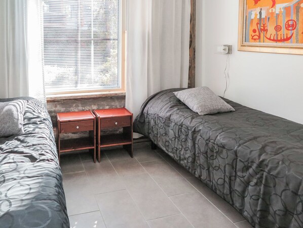 A bedroom with two single beds, a wooden nightstand, a window with blinds, and a wall-mounted power outlet.
