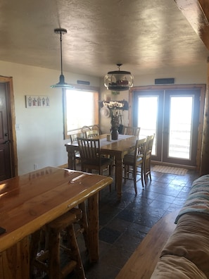 A rustic dining area with a wooden table and chairs, a hanging light fixture, and a sign above the door reading "The Gathering Place".