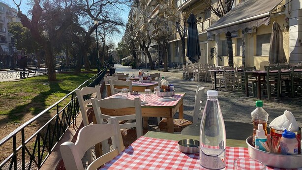 An outdoor café with tables and chairs, a bottle of water, condiments, and a view of a building with balconies. - Sunny Apartment (90m2) in Athens center (Athens)