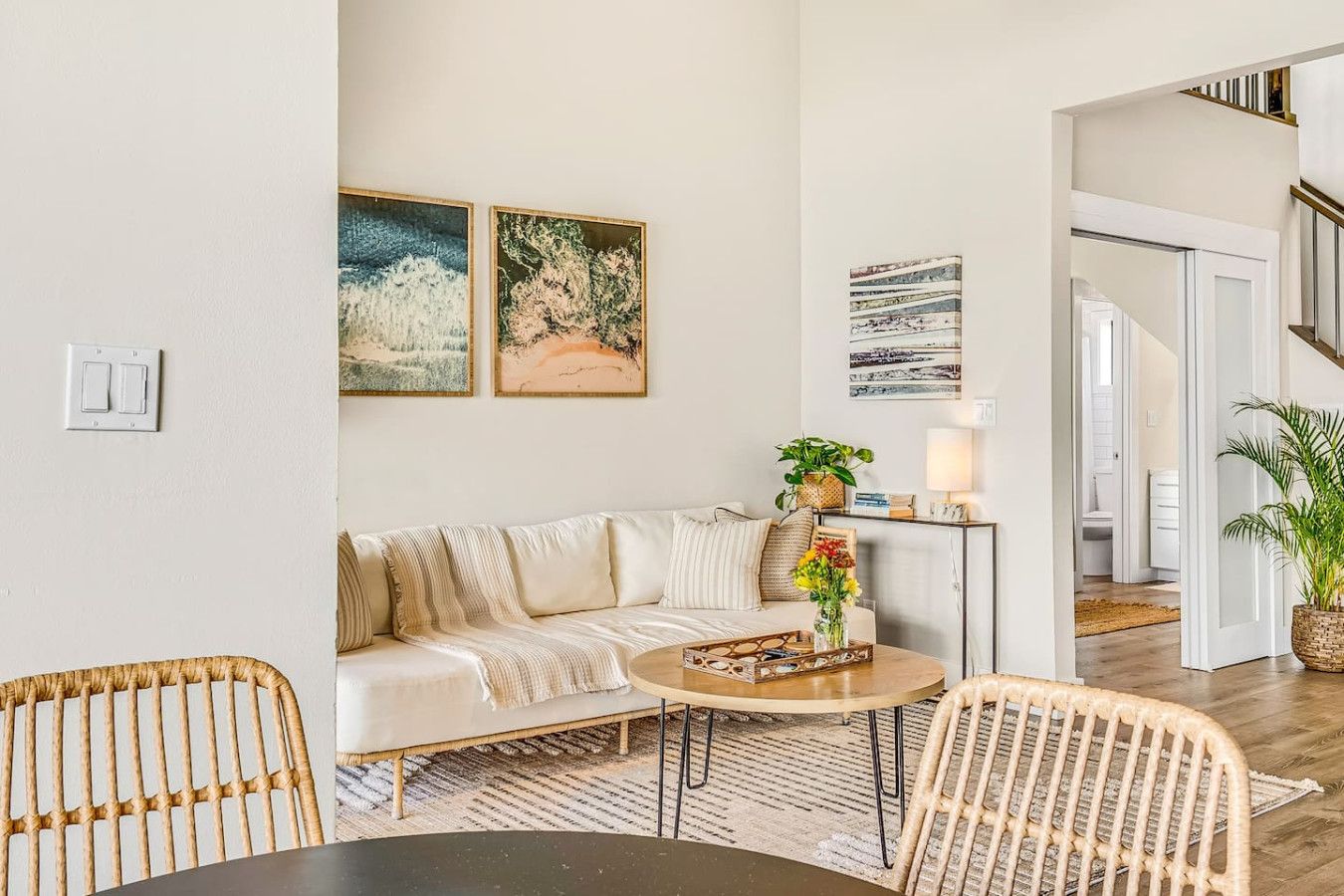 A modern living room with a sofa, a coffee table, and two wicker chairs. There are wall-mounted artworks and a potted plant on a side table.