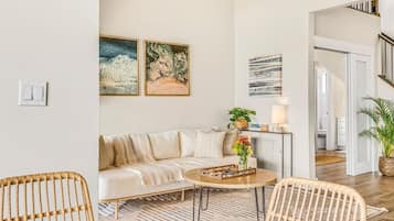 A modern living room with a sofa, a coffee table, and two wicker chairs. There are wall-mounted artworks and a potted plant on a side table.