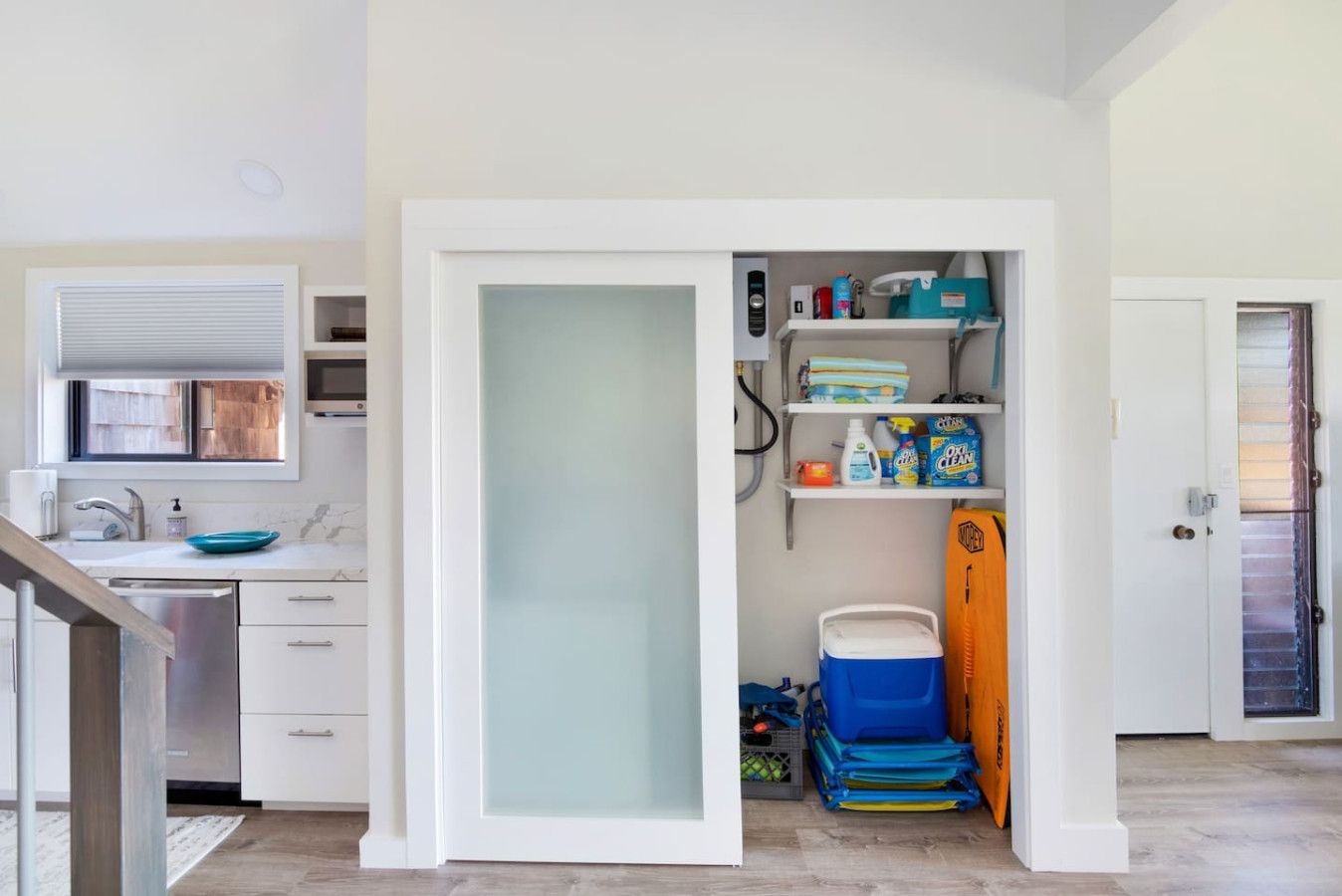 A compact kitchen with a built-in dishwasher, a window with blinds, and a storage area with cleaning supplies and a cooler.