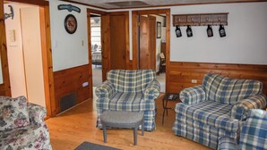 Una habitación acogedora con sofás de cuadros, un sillón floral y una pequeña mesa. Hay puertas de madera y un estante con botellas.