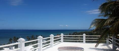 A terrace with a white railing, a round table, and benches, overlooking a coastal view with palm trees.