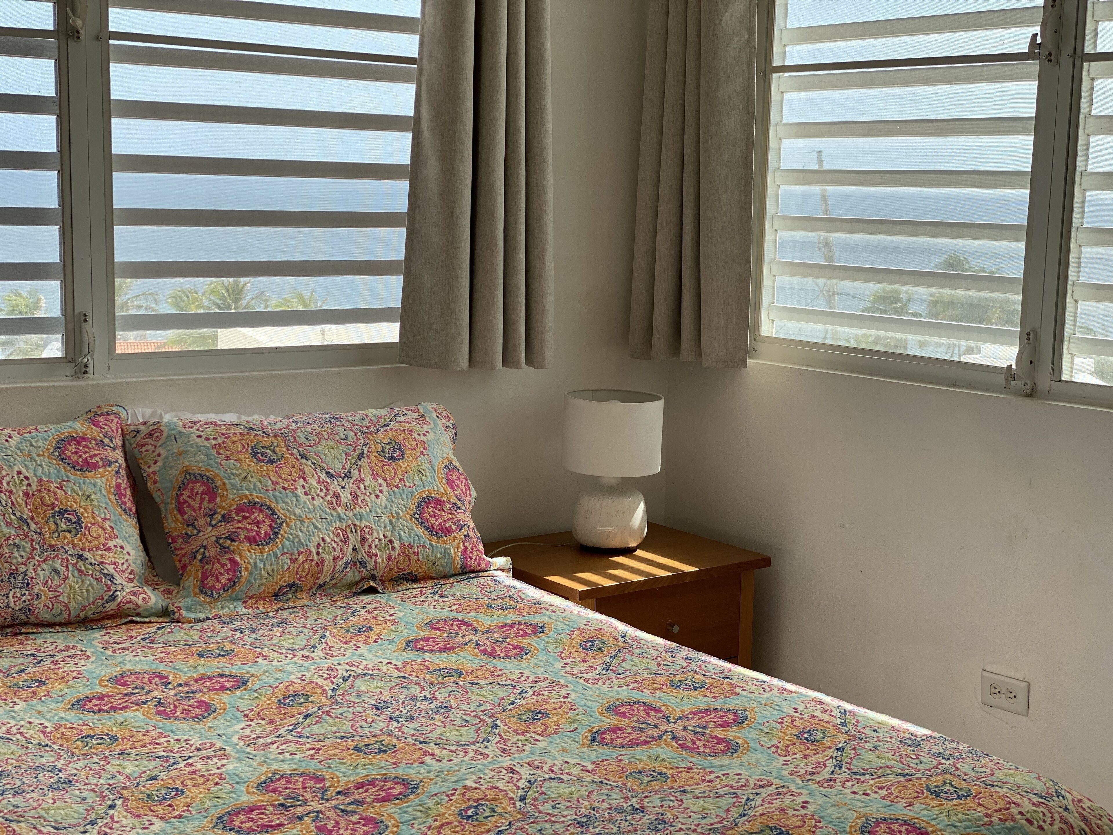 Un dormitorio con cama, dos almohadones estampados, una mesita de noche con lámpara y una ventana con cortinas.
