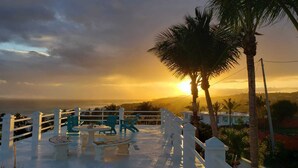 Eine Terrasse mit Tisch und Stühlen, die Blick auf einen Sonnenuntergang mit Palmen und eine entfernte Landschaft bietet.