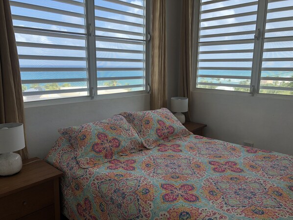 A bedroom with a bed, two bedside tables, a lamp, and a view of the sea through large windows with blinds. - Best Views in Vieques! (Vieques)