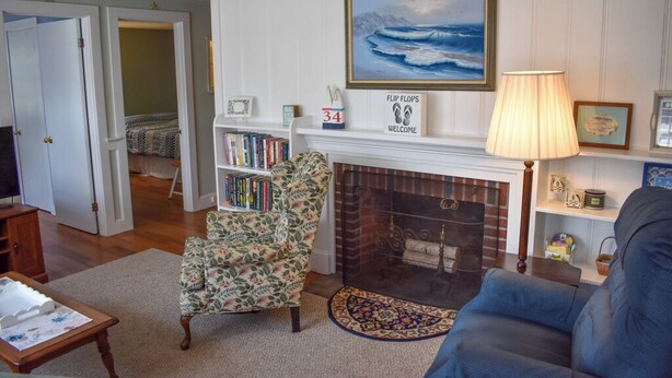 A living room with a fireplace, a bookshelf, a floral armchair, and a blue sofa. - Neat, clean and affordable 4 bedroom home with central air in West Dennis (West Dennis)