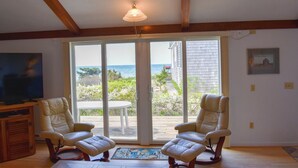 Una habitación con dos sillones de cuero, un televisor sobre un mueble de madera y un amplio ventanal corredero que da a un balcón con vista al océano.