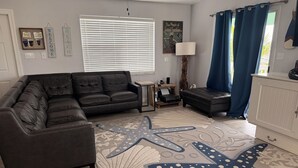 A living room with a sectional sofa, a small side table, a lamp, a cabinet, and a large window with blinds.