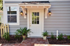 A house with a white front door, two wall-mounted lanterns, a picket fence, and potted plants.