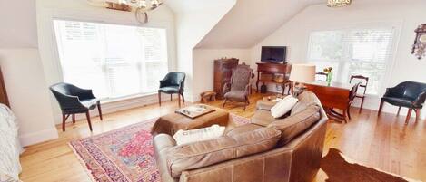 A spacious living room with a brown leather sofa, a wooden coffee table, and a patterned rug. There are two black chairs, a wooden sideboard, and a television. The room has a high ceiling with a fan and a chandelier.