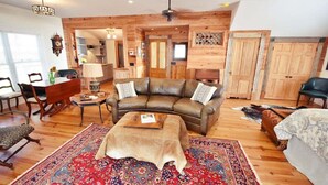 Una sala de estar acogedora con un sofá de cuero, paredes de madera, un tapete rojo estampado y una zona de comedor con mesa y sillas.