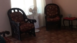 Un salon avec deux fauteuils à motifs, un ventilateur sur pied, une petite table recouverte d’une nappe rouge et un sol carrelé.