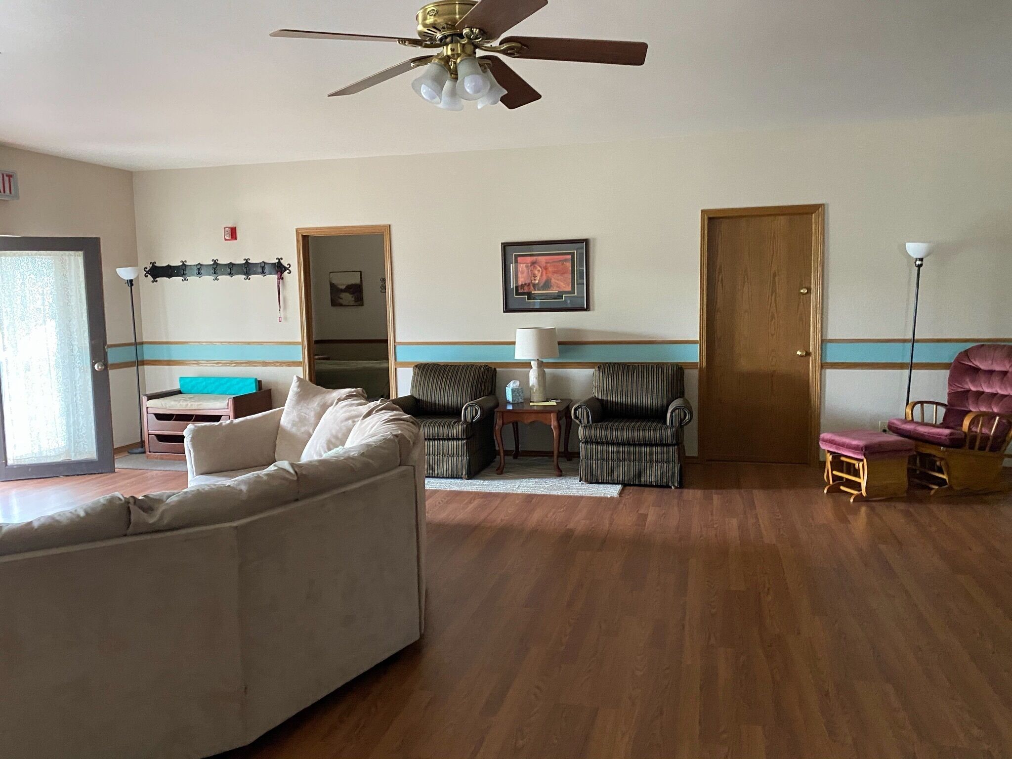 A living room with a ceiling fan, a sofa, two armchairs, a wooden floor, and a door.