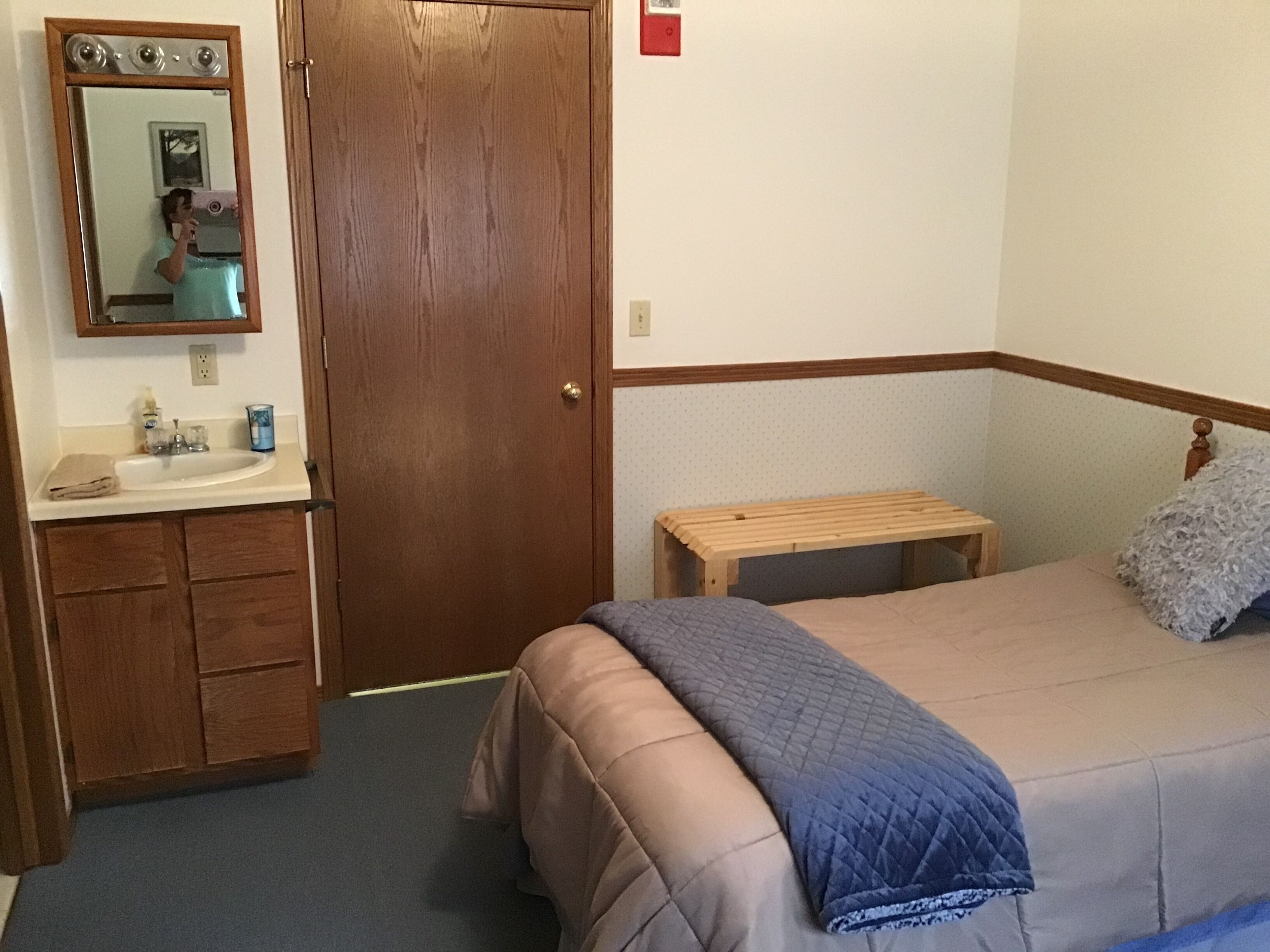 Um quarto com porta de madeira, uma pequena mesa de cabeceira, uma cama com manta acolchoada, um espelho sobre a pia e uma toalha no balcão.