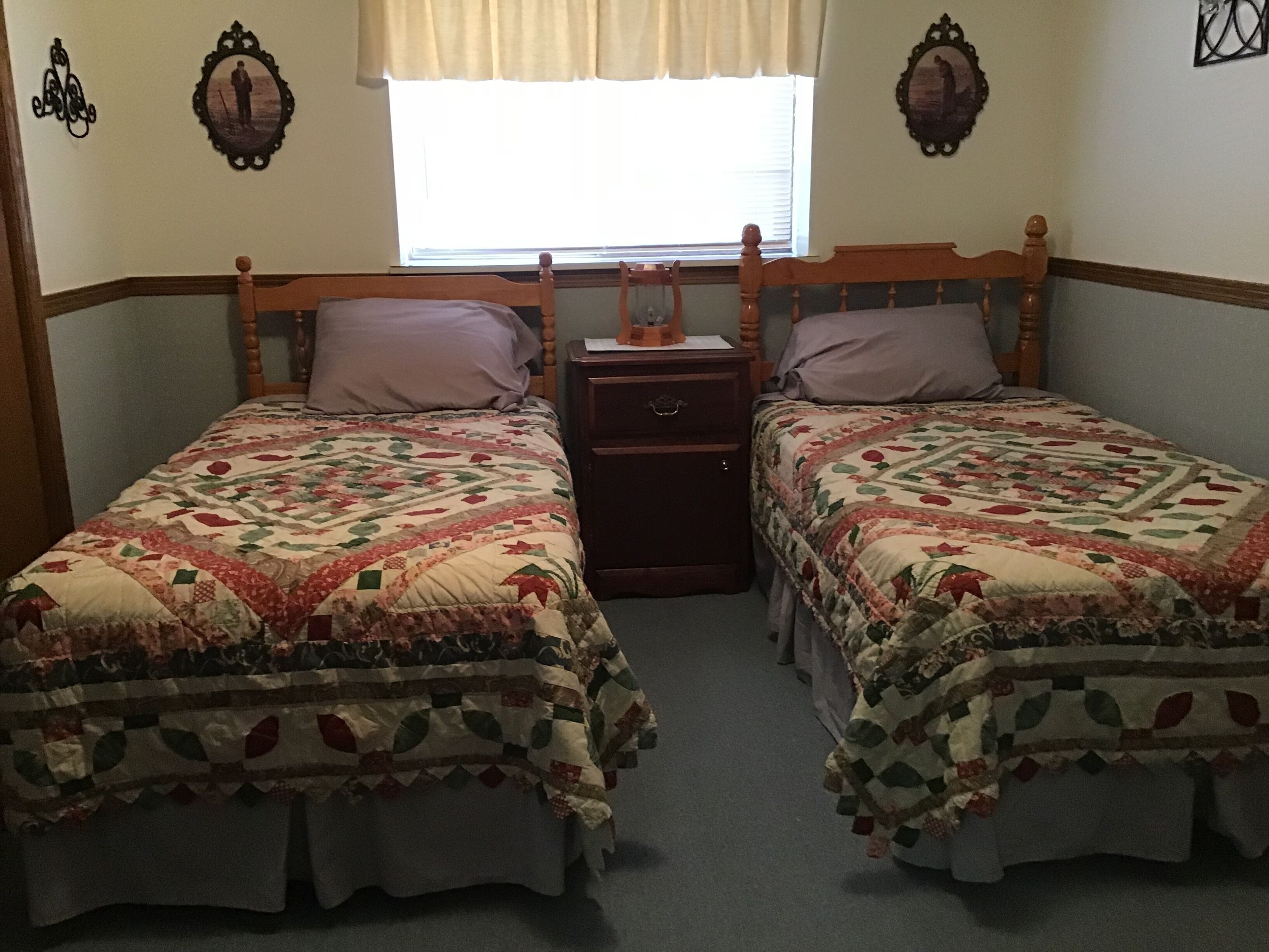 A room with two wooden beds, each with a floral quilt. There is a nightstand between the beds, and a window with curtains.
