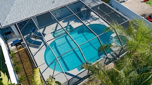 Vue aérienne d’une piscine dotée d’une structure en verre, entourée de palmiers et d’un espace patio aménagé avec des chaises et des tables.