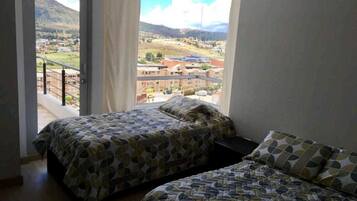 A bedroom with two beds, a large window with a view of a town and mountains, and a balcony.