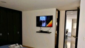 A hotel room with a bed, a TV mounted on the wall, and a door leading to another room. - Aparta Suite Ventus (Tunja)