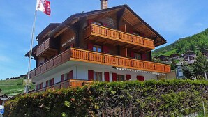 A two-story wooden chalet with balconies, a flag, and a mountainous backdrop. - Bergfink Obergeschoss by Interhome (Grächen)