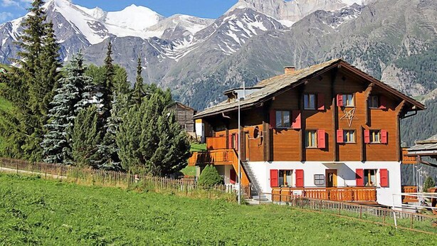 A wooden chalet with a sloped roof, multiple windows, and a staircase leading to the entrance. It is surrounded by a wooden fence and a lush green field. - Bergfink Obergeschoss by Interhome (Grächen)