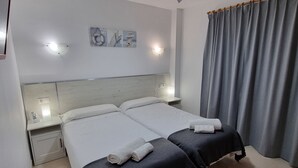 Habitación de hotel con dos camas, una mesita de noche con lámpara, un cuadro colgado en la pared y una cortina gris.